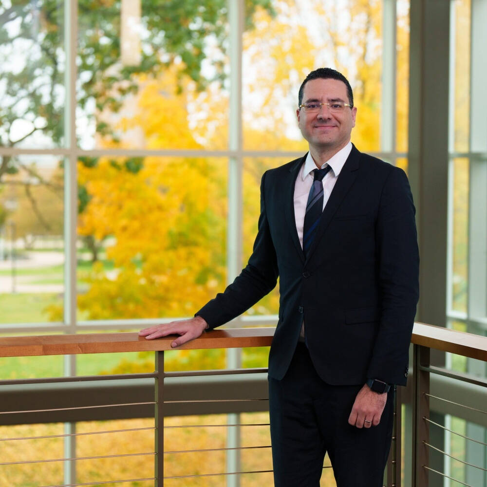 Dean Marouane Kessentini stands in a corner with large windows with visible yellow leaves in the background.